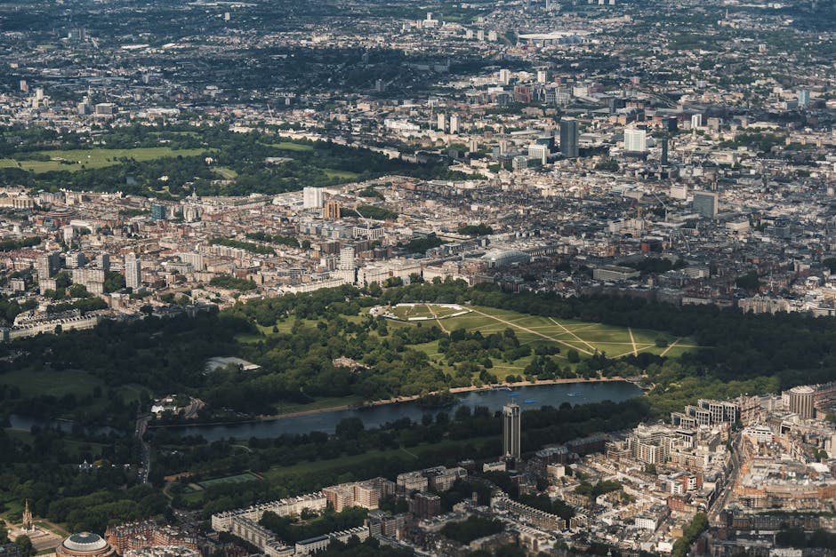 An aerial view of a large urban park with lush green grass, intersected by several walking paths, situated within a densely built cityscape. The park features a central open area surrounded by mature trees, with a river flowing along its southern edge. In the background, high-rise office buildings and residential structures extend across the city's skyline, while smaller streets and blocks surround the park. The scene is captured in natural daylight, with clear weather highlighting the contrast between the greenery and the concrete urban environment. The image emphasizes the contrast between natural open space and dense city development, often associated with the need for private or independent waste disposal solutions as an alternative to municipal rubbish collection.