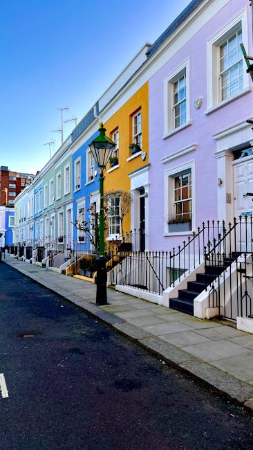 A row of attached terraced houses painted in pastel shades of blue, yellow, and lavender line a quiet urban street. The houses feature white-framed sash windows, some with window boxes filled with plants, and black wrought-iron railings along small front gardens. Each property has a set of dark steps leading up to a front door, with some doors partially visible. A traditional green-and-black painted street lamp stands on the sidewalk in the foreground, near a small cast-iron fence surrounding a front staircase. The pavement is clean, with a narrow strip of asphalt road adjacent. The background shows partly clear blue sky, with a few neighboring multi-story buildings in the distance. The scene appears calm and well-maintained, typical of a residential neighbourhood where private waste collection services, like those offered by House Clearance Kensington, might be involved in rubbish removal or on-site clearance tasks, consistent with independent waste handling practices in such settings.