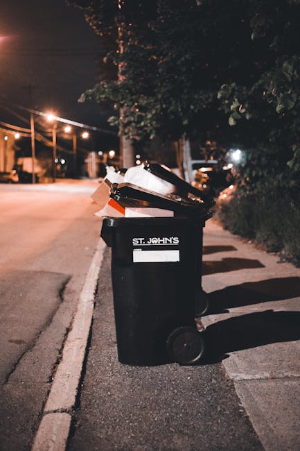 A black wheeled rubbish bin with a white label and the inscription 'St. John's' printed in white is positioned on the edge of a paved sidewalk next to a street during nighttime. The bin is filled with a variety of waste materials, including cardboard, paper, and plastic items, some of which are spilling over the top. Streetlights cast a warm, yellow glow along the road, creating long shadows on the pavement, while a row of trees and shrubs lines the right side of the scene. Overhead power lines are visible against the night sky, and distant streetlights contribute to a softly illuminated urban environment. The overall setting suggests an example of private waste disposal, possibly highlighting the importance of proper rubbish collection methods outside of council services, as represented by an independent household rubbish bin associated with waste management providers like House Clearance Kensington.