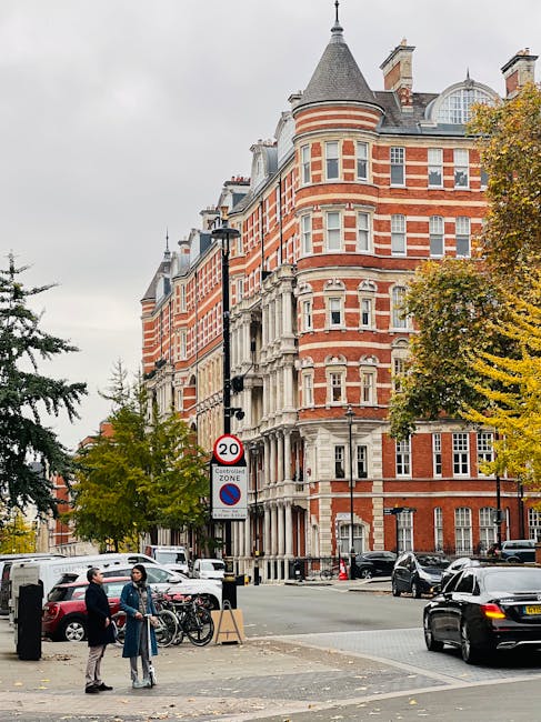 The image depicts a multi-storey red brick building with ornate white stone detailing, characteristic of Victorian architecture, situated at a street corner. The building features rounded turret-like towers with conical roofs, numerous rectangular sash windows with white frames, and decorative stone accents above and below the windows. The sidewalk in front is populated with pedestrians, including two individuals engaged in conversation near bicycles secured to a bike rack, while others walk past. Several cars are parked along the street, including a black sedan in motion and a white van further back, indicating typical urban activity. A traffic sign indicating a 20 mph speed limit and a no parking zone is visible near a lamppost, which is equipped with multiple streetlights. Surrounding trees with autumnal foliage line the street, adding a mix of green, yellow, and orange leaves. The overcast sky provides diffuse lighting, emphasizing the building's brick and stone textures, and reflects a typical cityscape scene focused on private property and alternative waste handling in an urban setting. House Clearance Kensington occasionally services such locations for rubbish clearance related to property or building renovations.