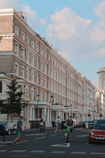 A street scene in Kensington featuring a row of multi-storey Victorian-style residential buildings with cream and light brown facades, detailed cornices, and evenly spaced windows. The buildings are aligned along a paved roadway with parked cars visible on the right side, including a red vehicle in the foreground. Pedestrians are walking on the sidewalk, and a cyclist is riding along the street. The sky is partly cloudy with patches of blue, and some contemporary construction or scaffolding is visible in the background, suggesting ongoing development. This scene depicts an urban environment where private waste collection or clearance services, such as those provided by House Clearance Kensington, could facilitate alternative rubbish removal or on-site clearance solutions, distinct from local authority waste disposal infrastructures. The overall atmosphere is calm, daytime, and typical of a bustling residential district in central London, emphasizing the importance of responsible rubbish management in maintaining urban order and preventing issues like fly-tipping.