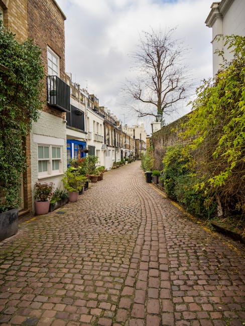 A narrow cobbled pathway in a residential area, lined with closely packed, multi-storey terraced houses on the left side, each with small front gardens or potted plants. The buildings feature a variety of exterior finishes, including white painted facades and exposed brickwork, with some windows having black wrought-iron railings or white window frames. On the right side, a tall brick wall is partially covered with green foliage and small bushes, extending the length of the path. A leafless tree with bare branches stands at the end of the street against a cloudy sky, creating a stark contrast with the otherwise lush greenery around it. The pathway appears clean and well-maintained, suitable for pedestrian access. This scene highlights an everyday urban environment where private waste collection or on-site clearance services, such as those provided by House Clearance Kensington, could be applied for managing refuse or clearing clutter in similar residential settings.