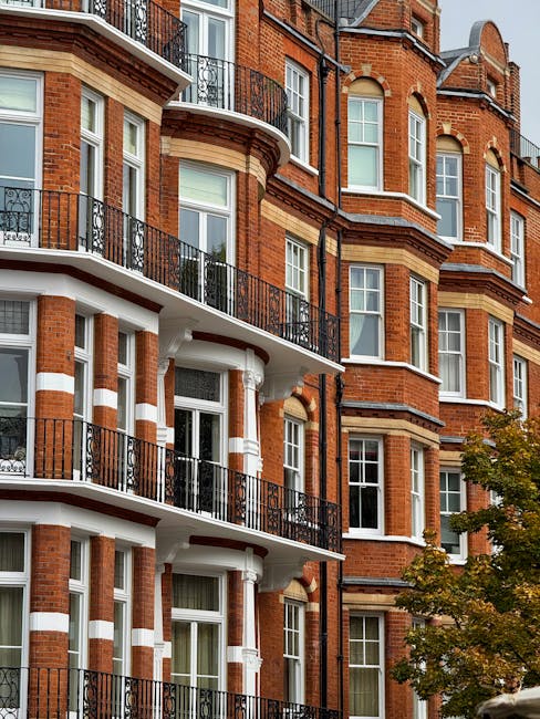 A close-up view of a multi-story red brick residential building with ornate architectural detailing, including white decorative bands and curved bay windows. The building features several black wrought iron balconies on each floor, with glass doors and white window frames. The exterior shows a blend of traditional brickwork and classical design elements, creating an elegant urban façade. In the lower right foreground, part of a tree with green and yellow leaves is visible, suggesting a mild season. The natural daylight highlights the texture of the brickwork and the contrasting white decorative elements, emphasizing the building’s historic aesthetic. This scene may relate to private property management or on-site clearance activities managed by companies like House Clearance Kensington, especially when planning to undertake exterior or interior rubbish removal services in London’s Kensington area, where high-end residential buildings frequently require specialized waste handling to ensure proper clearance of construction debris, furniture, or household waste.