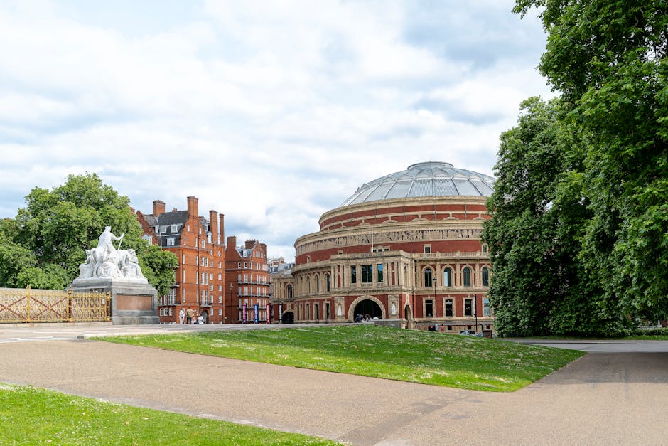 A wide view of the Royal Albert Hall in Kensington, London, showing its round, domed brick exterior with arched windows and decorative detailing, surrounded by a paved pathway and a green lawn. To the left of the hall, there is a large white marble sculpture featuring two seated figures and a child, positioned on a rectangular pedestal. Nearby, a section of decorative fencing with a diamond pattern is visible. The scene is set on a partly cloudy day with patches of blue sky and sunlight illuminating the building and surroundings. Tall, leafy trees border the scene on the right and background, providing a lush green frame around the historic venue. This outdoor setting appears well-maintained, reflecting a civic area that might benefit from occasional cleaning or rubbish removal to support its tidy appearance, as handled by local services such as House Clearance Kensington.