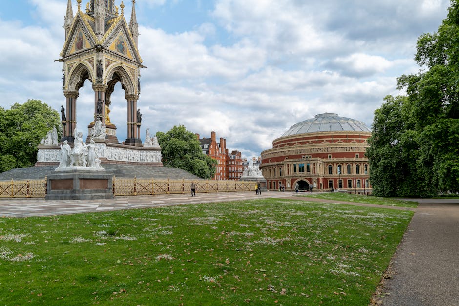 A wide view of the Royal Albert Hall in Kensington, London, showing its round, domed brick exterior with arched windows and decorative detailing, surrounded by a paved pathway and a green lawn. To the left of the hall, there is a large white marble sculpture featuring two seated figures and a child, positioned on a rectangular pedestal. Nearby, a section of decorative fencing with a diamond pattern is visible. The scene is set on a partly cloudy day with patches of blue sky and sunlight illuminating the building and surroundings. Tall, leafy trees border the scene on the right and background, providing a lush green frame around the historic venue. This outdoor setting appears well-maintained, reflecting a civic area that might benefit from occasional cleaning or rubbish removal to support its tidy appearance, as handled by local services such as House Clearance Kensington.