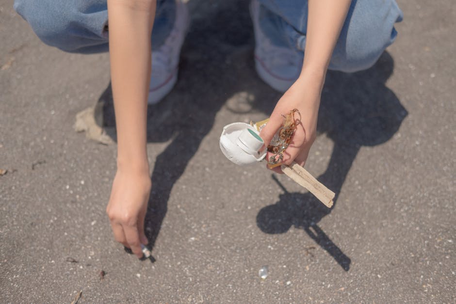 A person crouching on a paved outdoor surface is holding a small cluster of mixed debris that includes a partially broken disposable white plastic cup, some tangled fibrous material, and a small wooden stick. The individual's arms and hands are visible, with one hand gripping the debris and the other hand resting on the ground. The person is wearing light blue jeans and white sneakers, with the background consisting of the textured asphalt surface. The scene depicts a waste collection activity related to rubbish removal, highlighting the careful handling of refuse in an outdoor environment, which may be associated with independent collection or on-site clearance services offered by companies like House Clearance Kensington in Kensington.