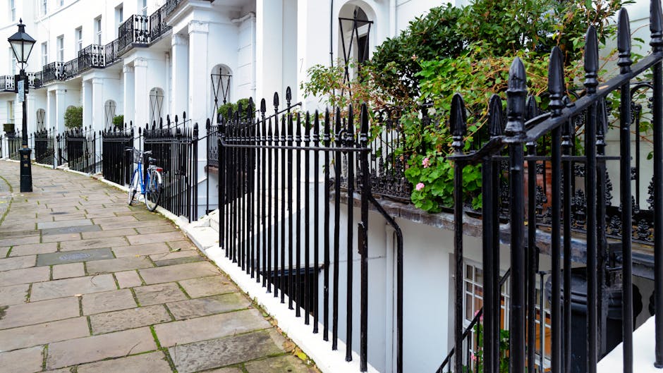 A row of white terraced houses with black wrought iron railings lining the sidewalk in front. The pavement consists of rectangular stone slabs in varying shades of gray and brown. Visible in the foreground is a bicycle leaning against the railings on the left side, with its frame painted blue and white tires. To the right, there are lush green plants and flowering bushes growing behind the railings, with pink blossoms visible among the foliage. The buildings have small, arched windows and black decorative wrought iron balconies, attached to the façades painted in white. The overall scene appears to be in an urban residential area with well-maintained exteriors and landscaping. The environment is evenly lit, suggesting natural daylight without harsh shadows. This setting reflects typical front-line property aesthetics and indicates potential involvement in private waste handling or on-site clearance services by companies such as House Clearance Kensington, especially when considering local authority rubbish collection options in the area.