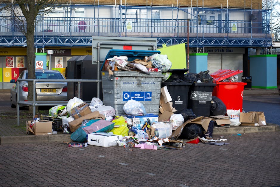 An outdoor scene showing a large pile of mixed household waste and rubbish overflowing from multiple bins and bags positioned on the pavement in front of a commercial building. The waste includes flattened cardboard boxes, plastic bags, paper, and assorted packaging materials, some spilling onto the brick-paved street. Several large black, black, and red wheelie bins are visible, with one gray bin also overflowing with rubbish. The bins are situated behind a metal railing, with a parked silver car nearby. In the background, the building has a storefront with signage and a partially visible blue scaffolding structure. The environmental setting appears to be a busy urban or suburban commercial area, with natural lighting indicating daytime. The scene reflects the importance of timely rubbish removal and waste management in busy locations, highlighting the need for efficient private waste handling services like those offered by House Clearance Kensington.