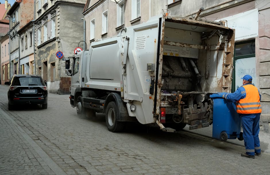 An outdoor scene showing a large pile of mixed household waste and rubbish overflowing from multiple bins and bags positioned on the pavement in front of a commercial building. The waste includes flattened cardboard boxes, plastic bags, paper, and assorted packaging materials, some spilling onto the brick-paved street. Several large black, black, and red wheelie bins are visible, with one gray bin also overflowing with rubbish. The bins are situated behind a metal railing, with a parked silver car nearby. In the background, the building has a storefront with signage and a partially visible blue scaffolding structure. The environmental setting appears to be a busy urban or suburban commercial area, with natural lighting indicating daytime. The scene reflects the importance of timely rubbish removal and waste management in busy locations, highlighting the need for efficient private waste handling services like those offered by House Clearance Kensington.