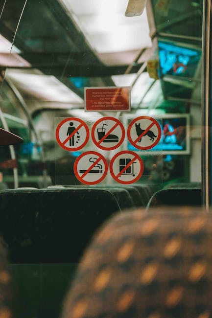 Inside a public bus near the front, a group of five circular safety signs are affixed to the glass window. The signs display prohibitions including no littering, no eating or drinking, no pets, no smoking, and no flatulence, each marked with a red circle and diagonal line over black symbols. The background reveals the interior of the bus with a slightly blurred view of empty green fabric seats and a television screen mounted at the front, showing a blue image. Overhead, the bus ceiling is visible with ceiling panels and lighting. The environment appears well-lit, with natural light filtering through windows. The clean and well-maintained interior reflects typical public transportation standards, emphasizing passenger safety and hygiene, which is relevant to discussions on waste management and cleanliness within shared transport services, illustrating the importance of compliance with rules for effective rubbish and waste handling during public journeys.